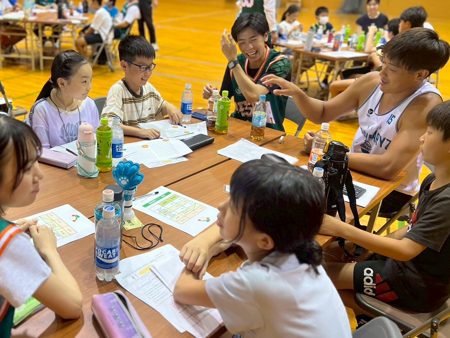 選手と子どもたちが一緒に机を囲みながら、金融教育プログラム「キッズマネーアカデミー」に参加している写真。